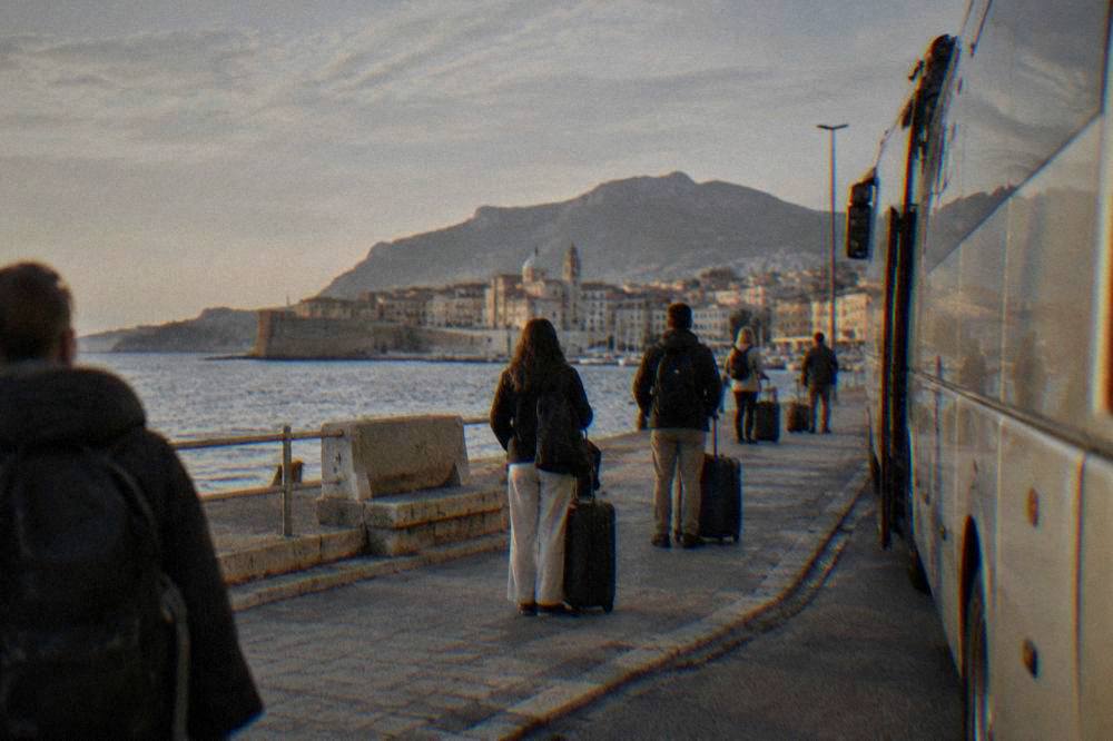 Passengers waiting at a coach stop with luggage in late afternoon light