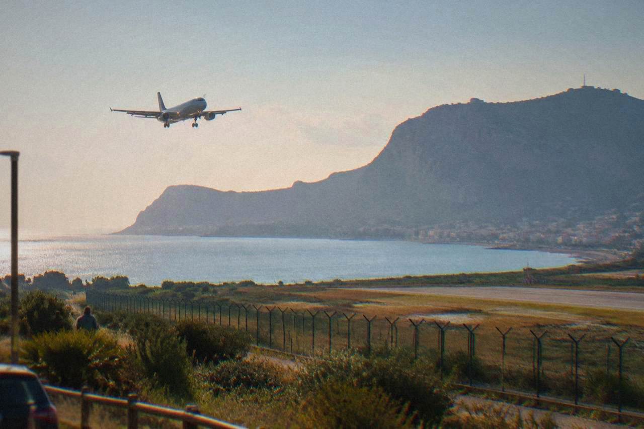 An airliner descending over a rocky coastline towards a sunlit Sicilian bay