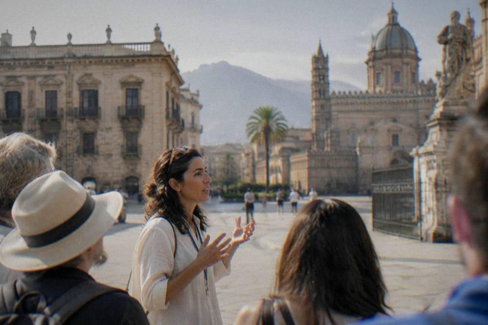 A guide addressing a small group of visitors in a sunlit Sicilian square
