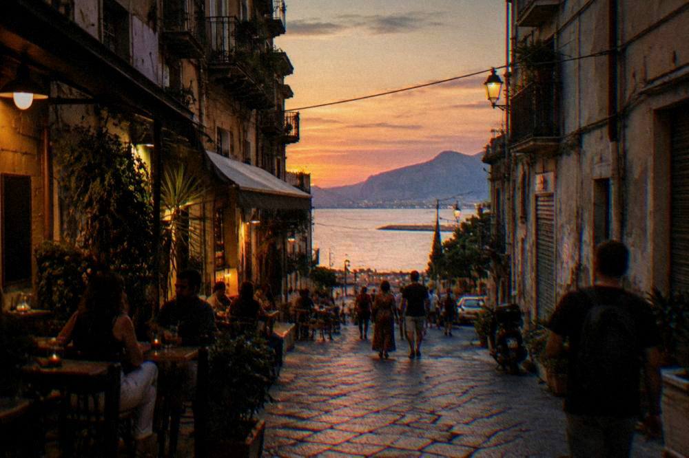 An evening street in the old centre of Palermo with lit bar fronts and people walking