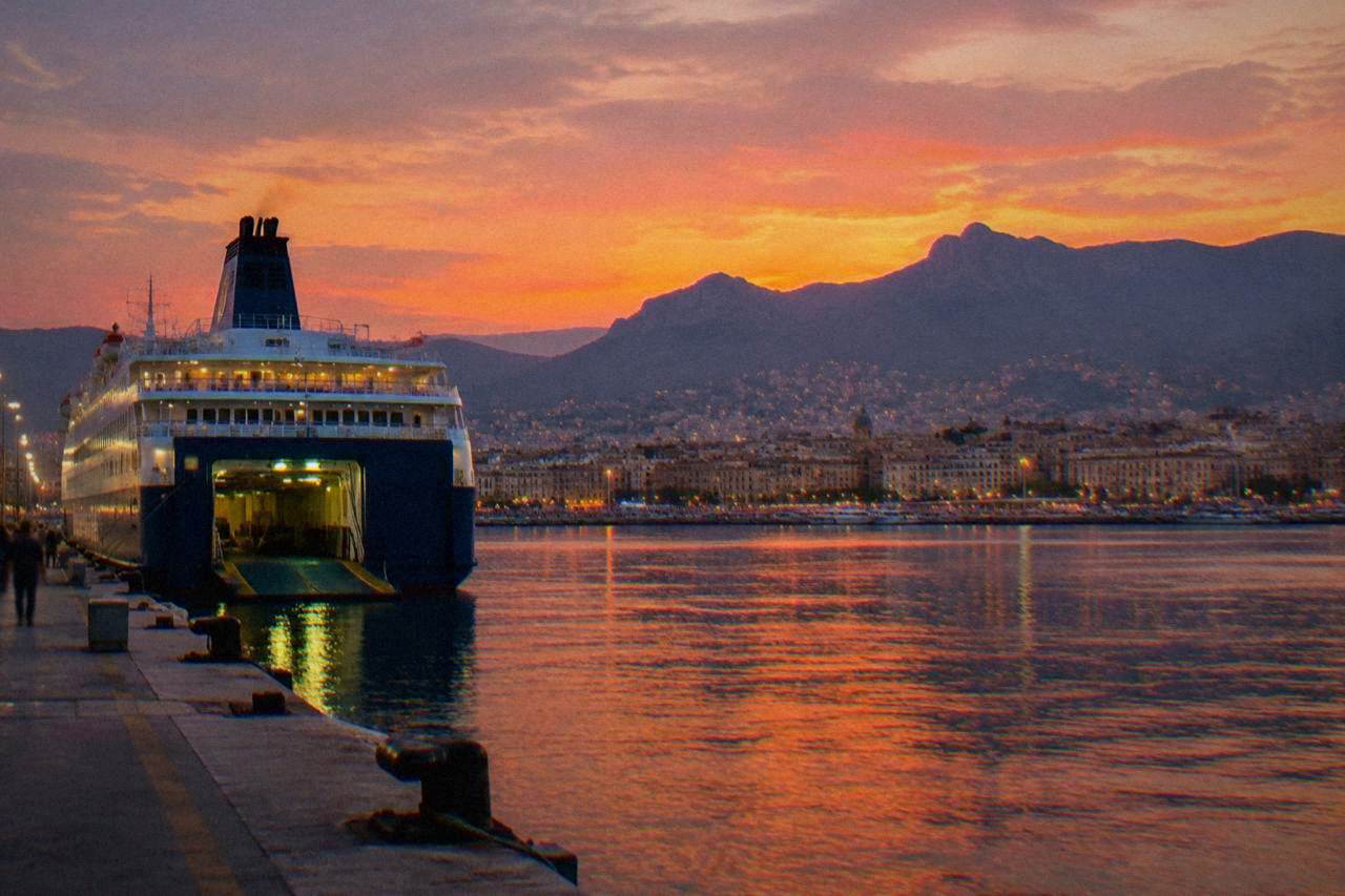 A large overnight ferry berthed at a Sicilian port at dusk, with the city lights of Palermo rising behind the harbour