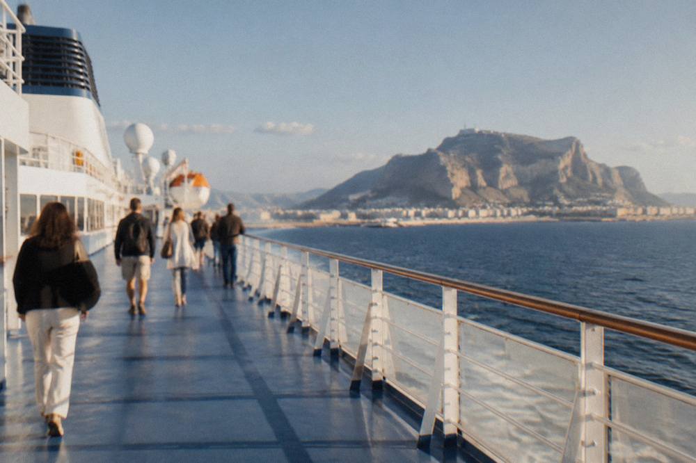 Passengers walking along the open deck of a ferry in the morning with the Sicilian coastline and a headland in view