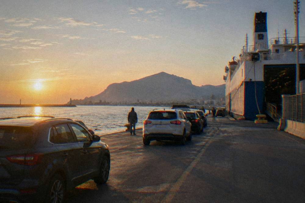 A ferry terminal quay in the early morning with a moored ship, waiting cars and a pale sunrise over the water