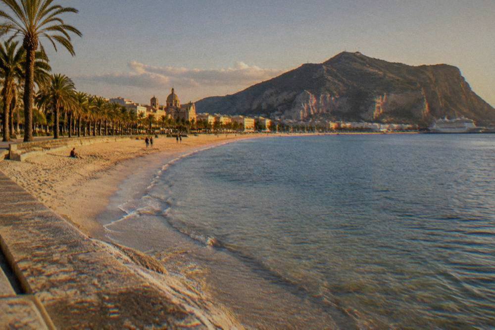 A wide sandy beach with pale sand, clear shallow water and a line of palm trees behind it at a Sicilian seaside town