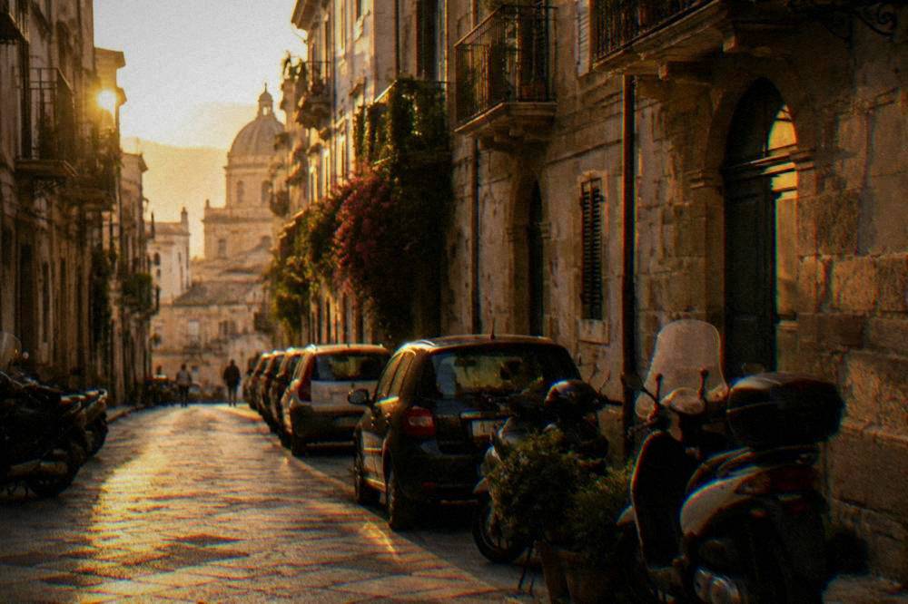 A narrow street in central Palermo with parked cars and scooters along the buildings