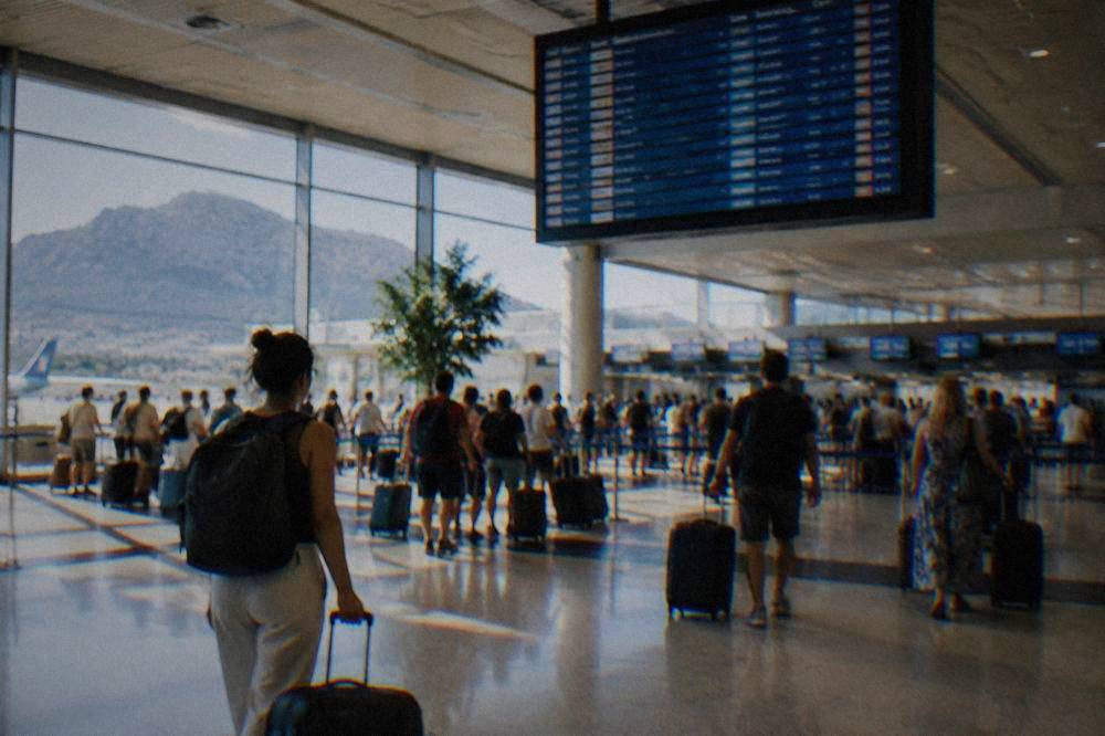 An airport departure board above a busy check-in hall with travellers pulling suitcases