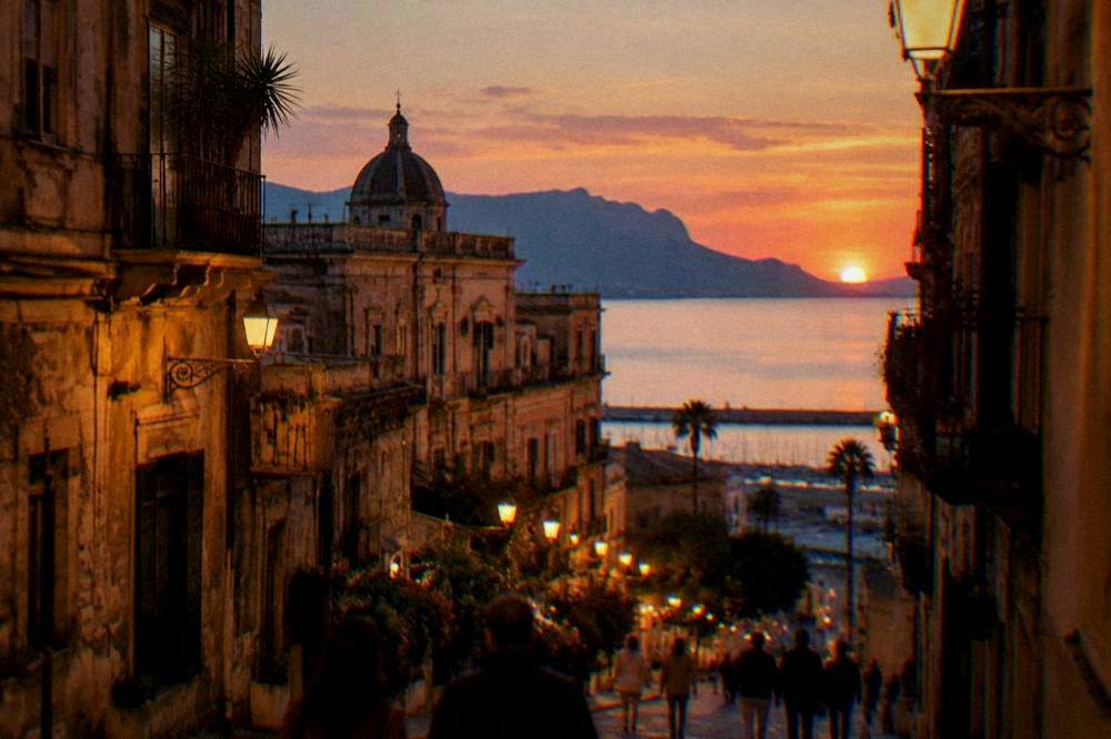 An evening street in a Sicilian city with warm lights on old buildings and people walking