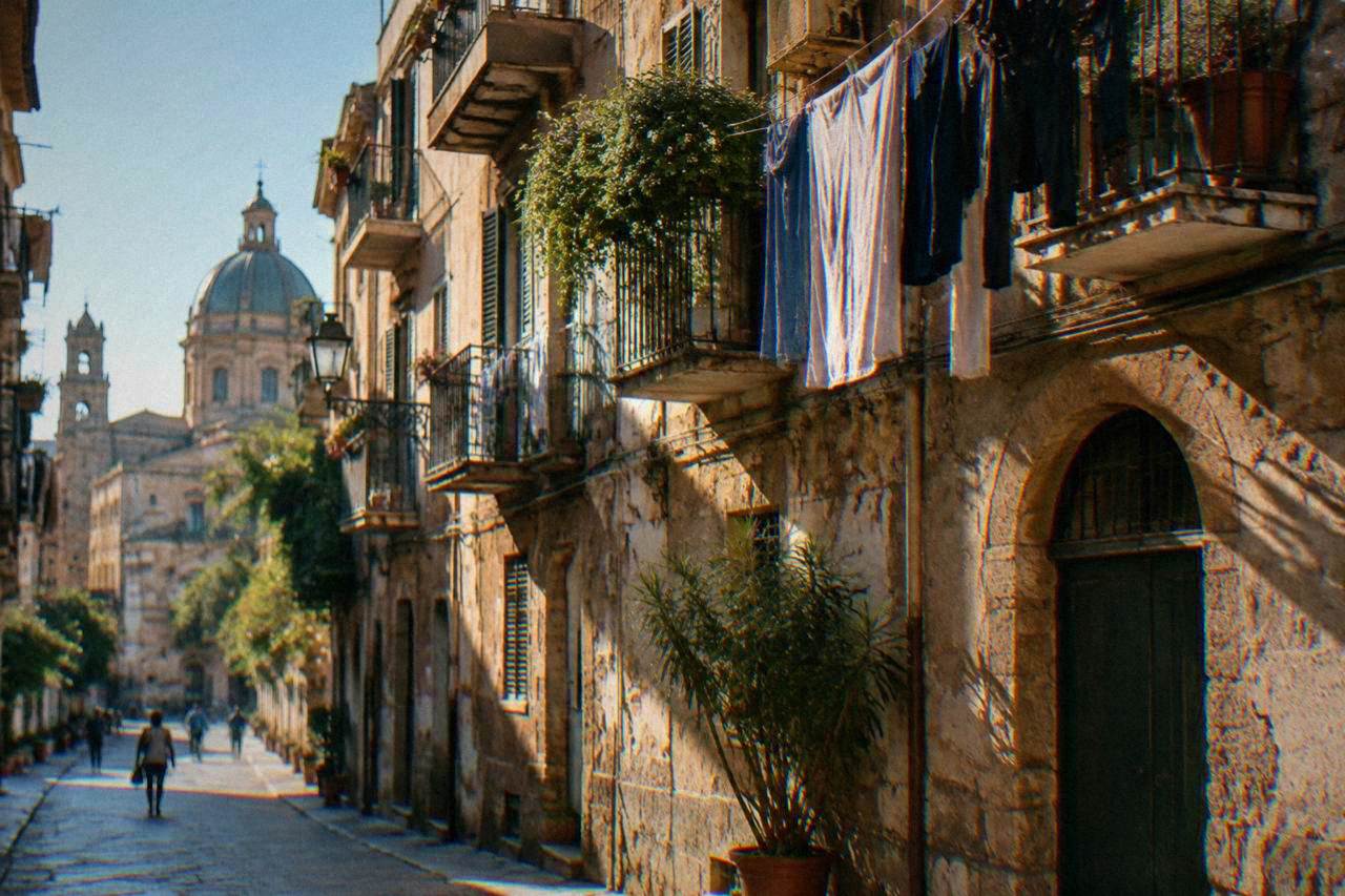A sunlit Palermo street with apartment balconies and laundry above an old stone facade