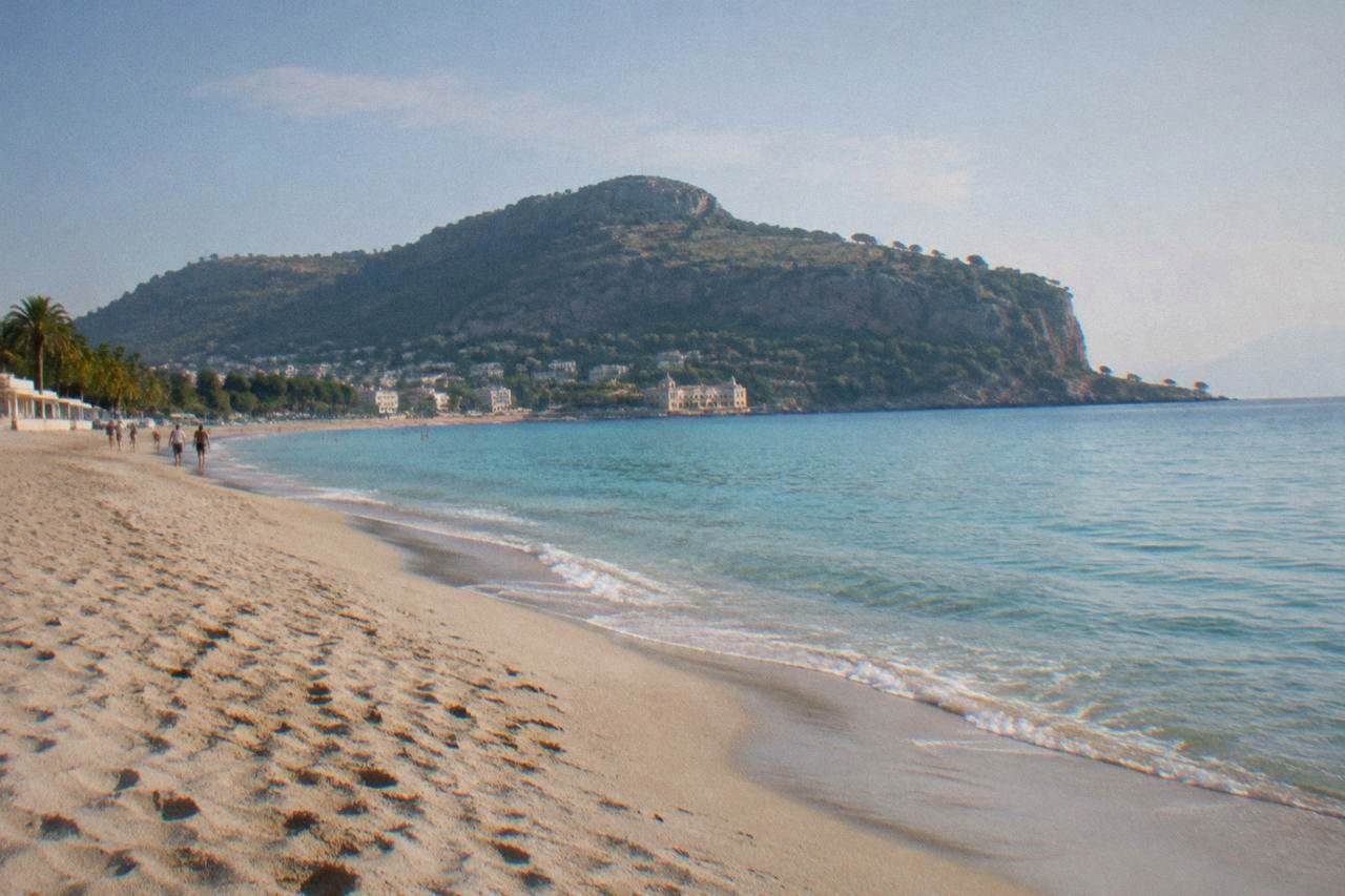 A wide sandy beach with calm turquoise water and a wooded headland in the background