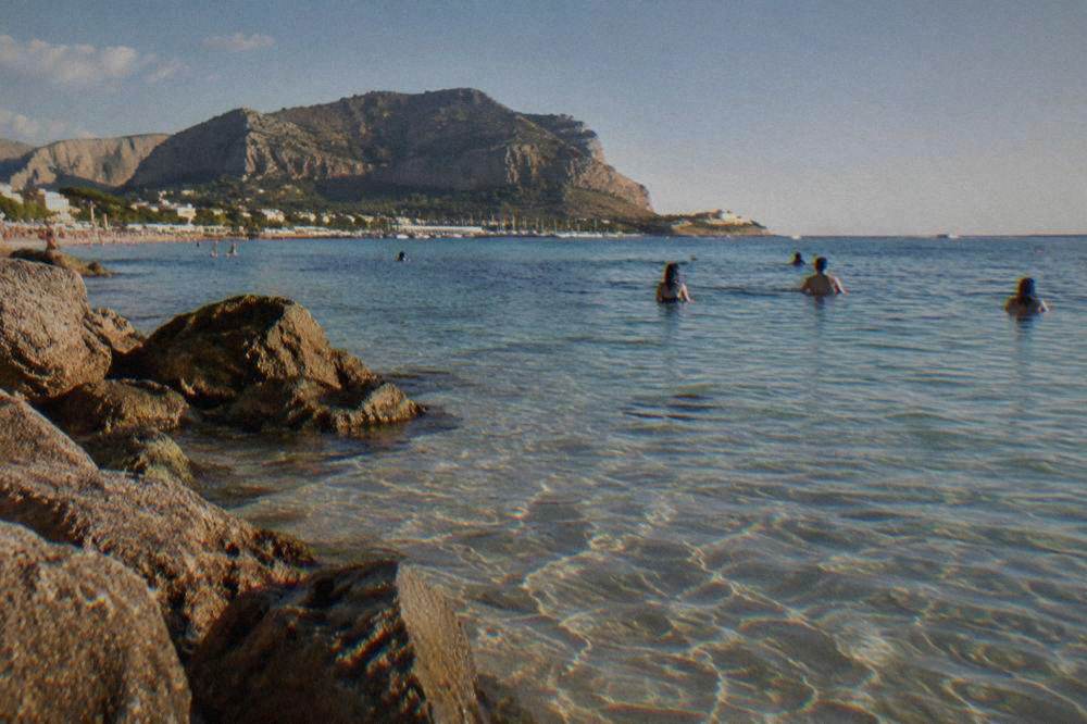 People swimming in clear shallow water near a rocky shoreline