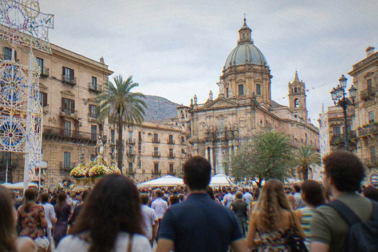 Crowds gathered in a sunlit Palermo square decorated for a summer festival, with historic facades and a church dome behind them