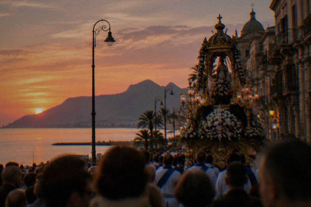 An ornate festival chariot decorated with flowers moving along a crowded historic street at dusk in Palermo