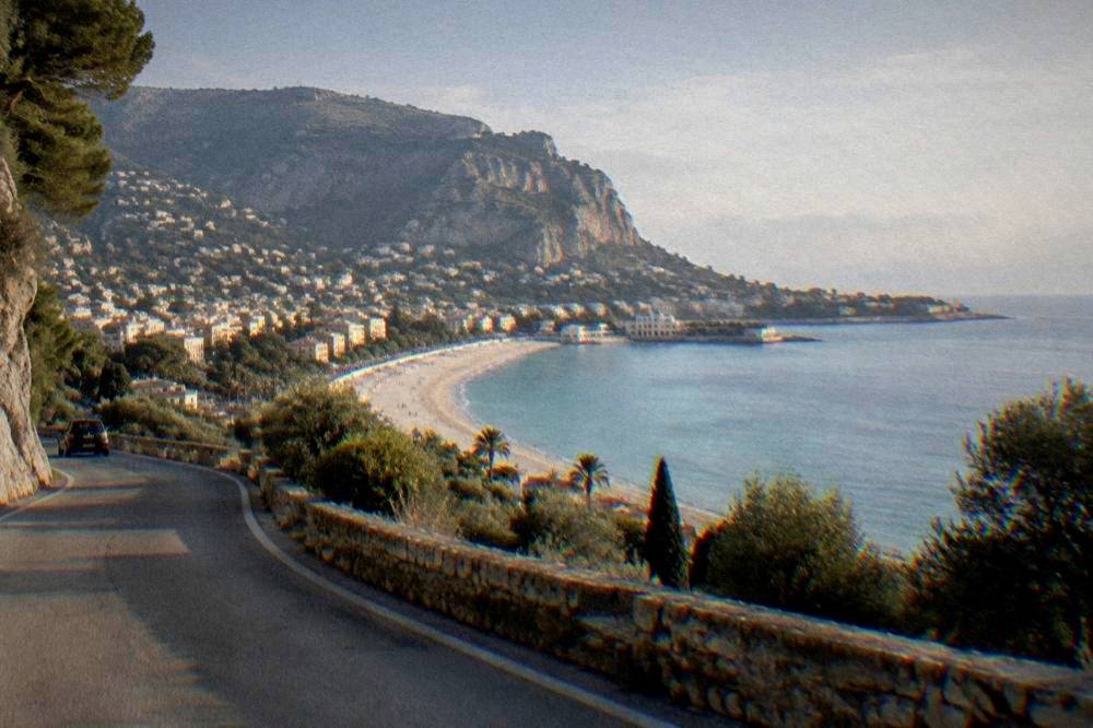 A hillside Sicilian town above the sea seen from a winding road, with a long sandy beach curving below