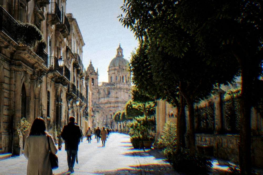 Visitors in coats walking a sunlit Palermo street lined with old buildings