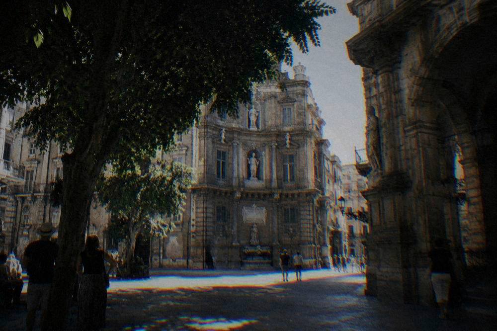 A historic palace courtyard in Palermo with arched stonework and visitors standing in the shade