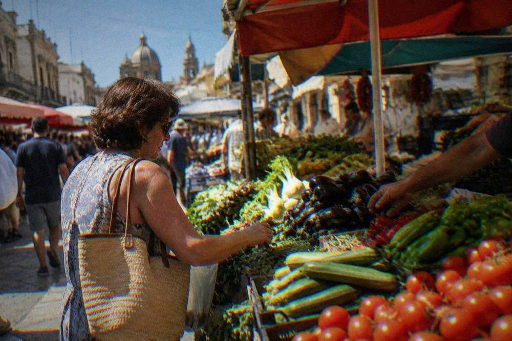 A shopper buying vegetables at a crowded Ballarò market stall in Palermo