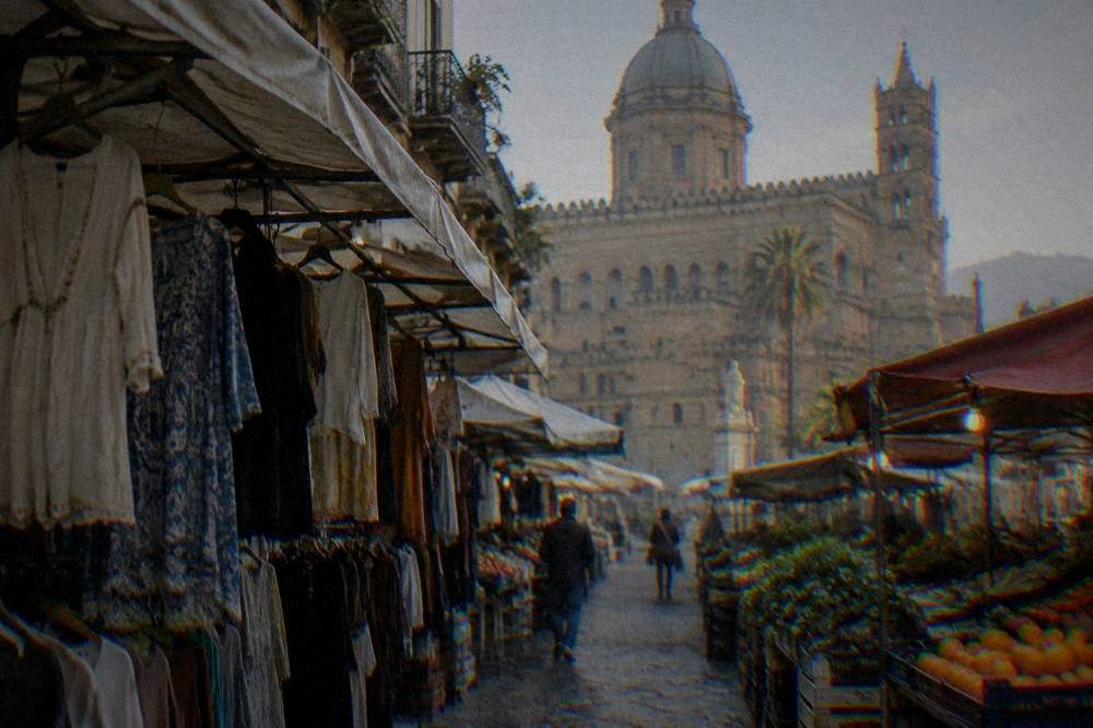 A covered market lane in the Capo district with hanging clothes and produce stalls near Palermo Cathedral