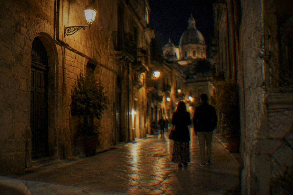 Two people walking down a warmly lit stone street at night
