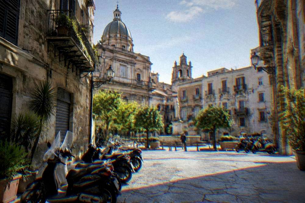 A quiet Palermo square in soft morning light with parked scooters and shuttered facades