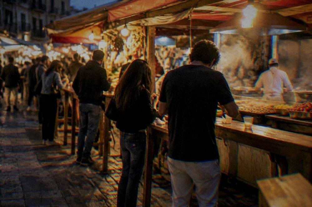 A Palermo street food counter at night with people eating standing at wooden benches