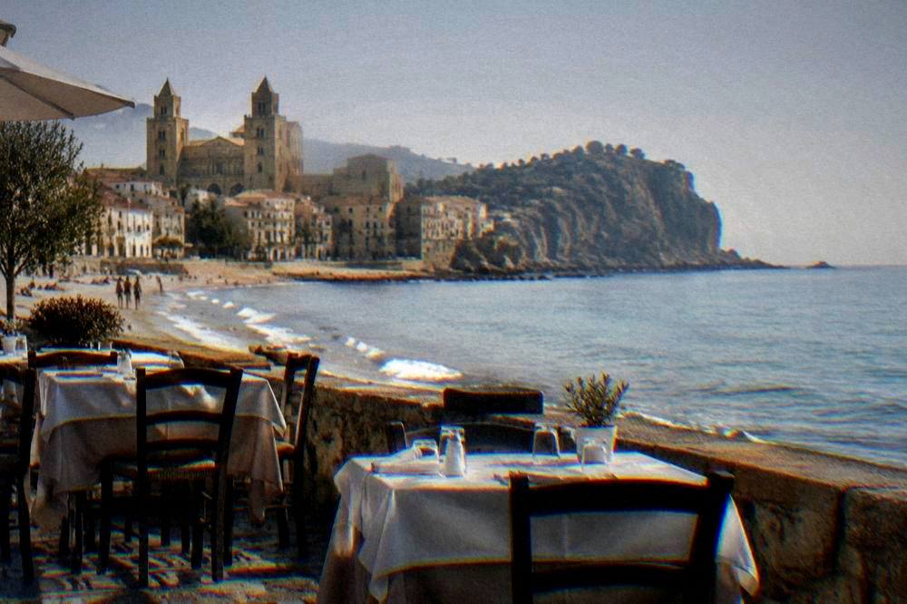 Seafront restaurant tables in Cefalu with the sandy beach and rocky headland behind