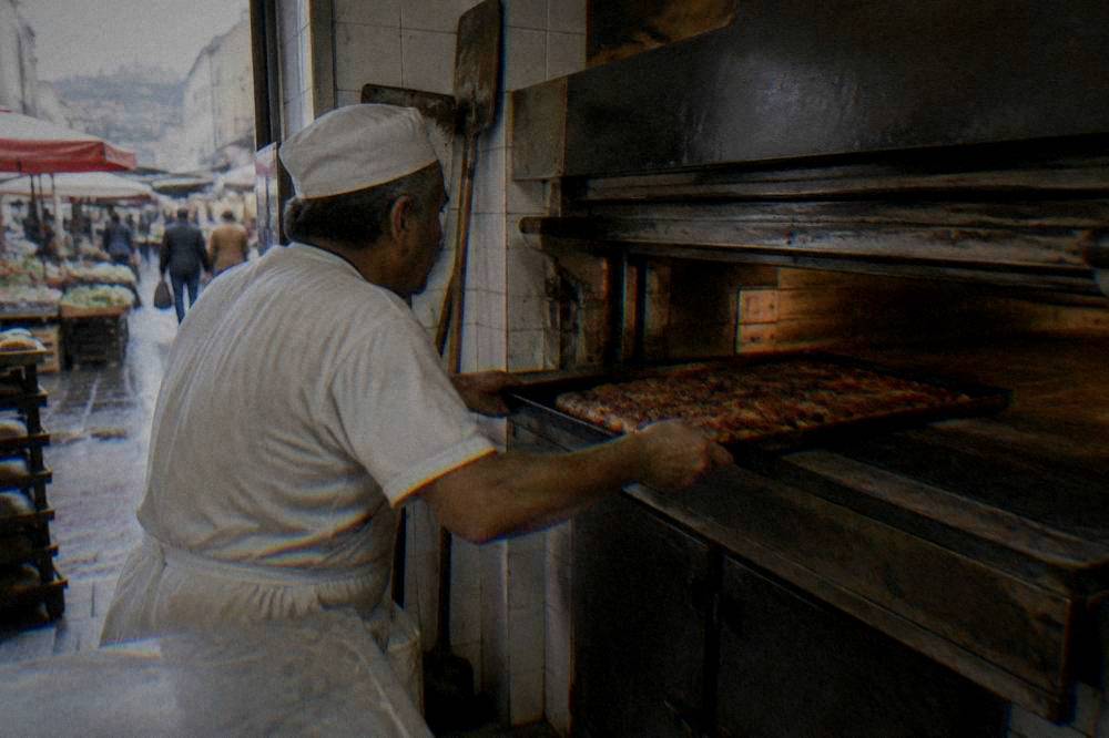 A baker sliding a tray of sfincione into a deck oven in a small Palermo bakery