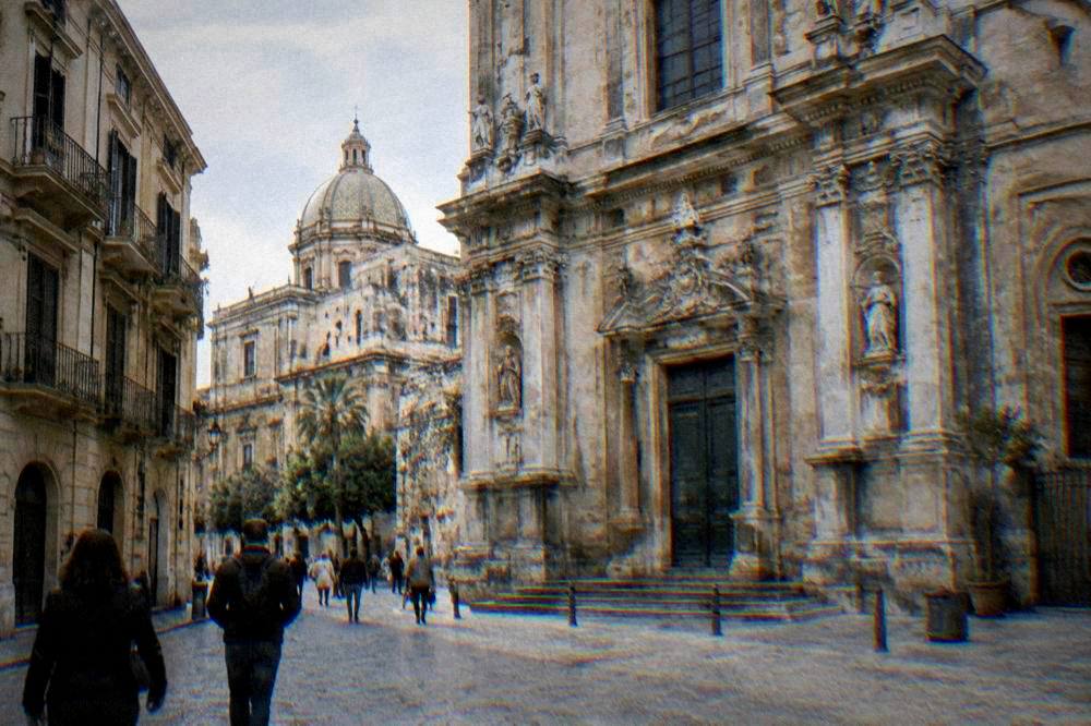 A Palermo street corner with a church facade and pedestrians in sunshine