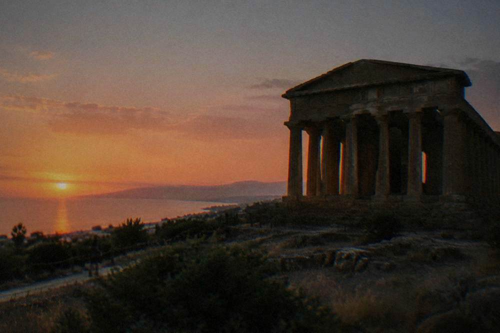 A stone Greek temple with columns silhouetted against an evening sky