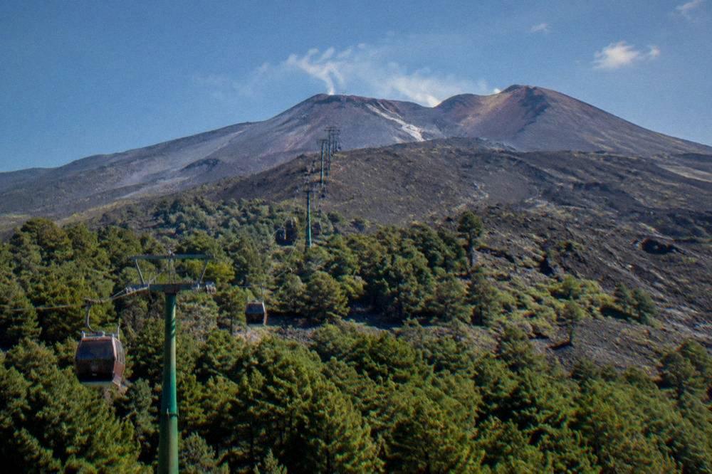 A rocky volcanic mountainside above forests with a cable car line climbing toward craters