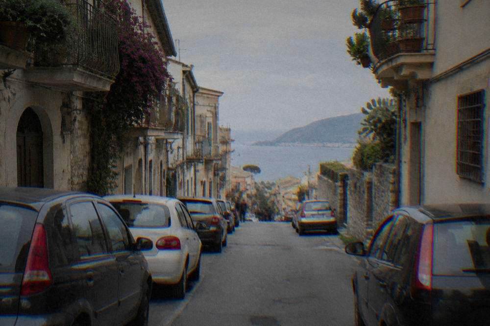A narrow Sicilian street with parked cars on both sides and old buildings above