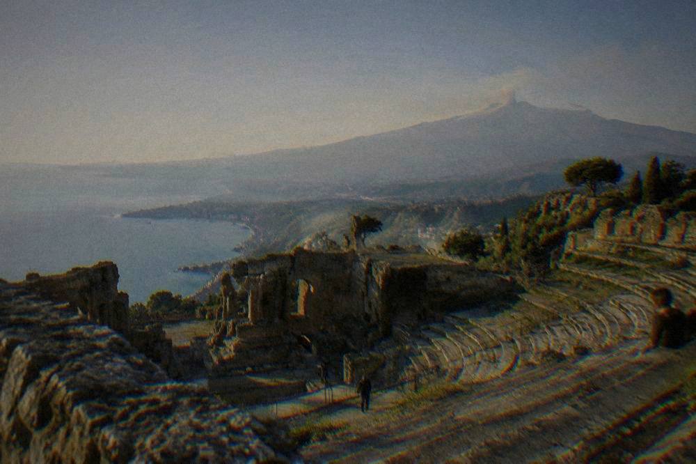 An ancient stone theatre seating row looking out over a coastline and a distant volcano