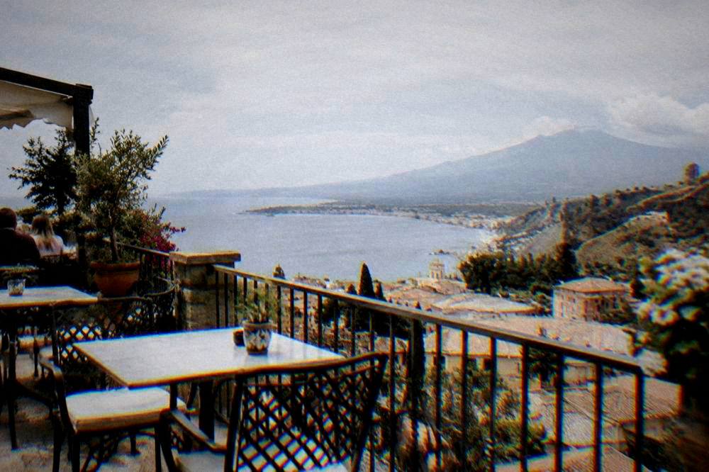 A sunny terrace with tables and chairs overlooking rooftops and a wide bay