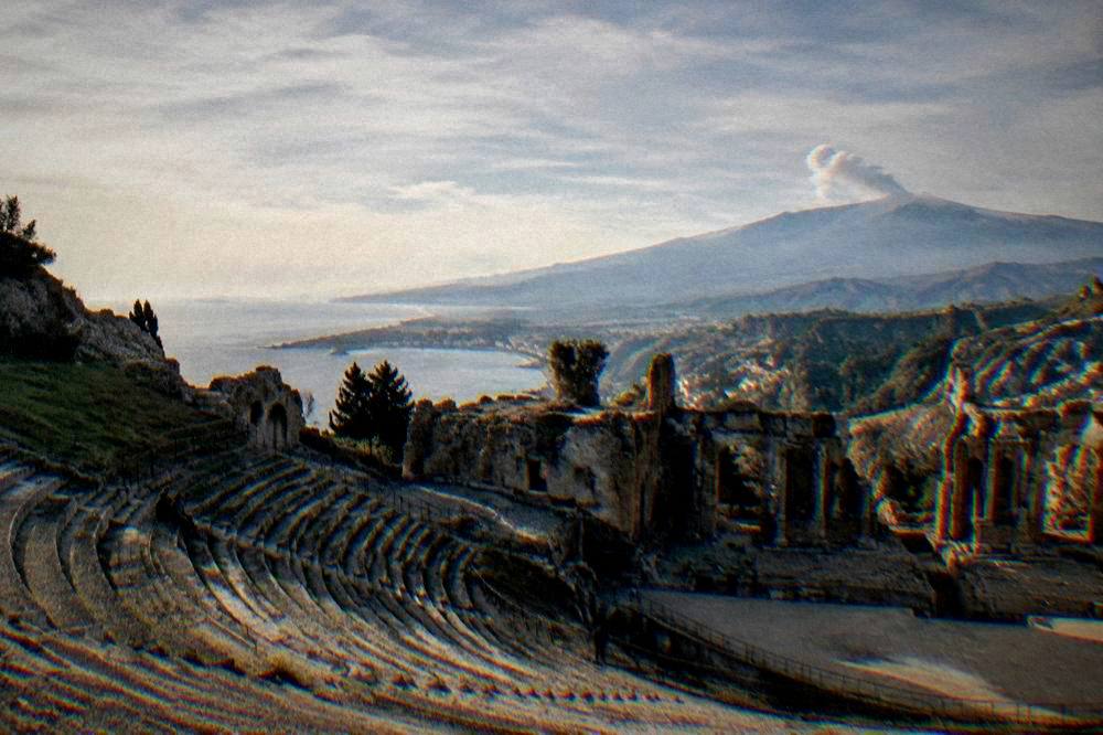 Ancient theatre seating facing the sea and a smoking volcano in the distance