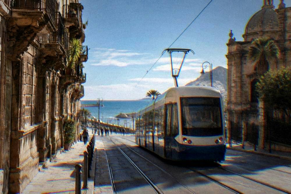 A tram moving along a Palermo street with the sea visible in the distance