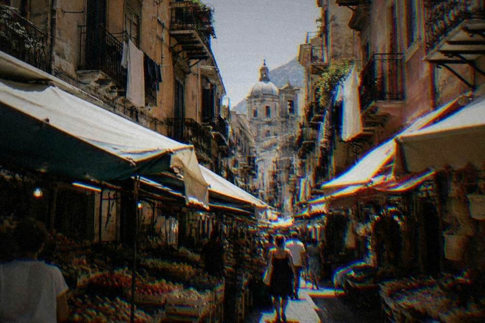 A narrow lane of faded ochre buildings with balconies, laundry lines and market stalls under awnings