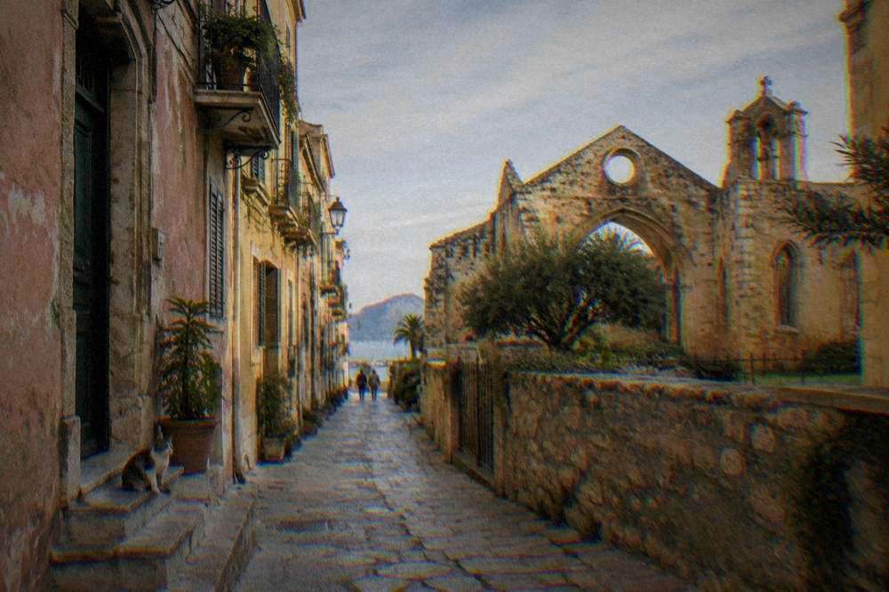 A quiet lane of faded pink palazzi beside a roofless old church with a cat on a step