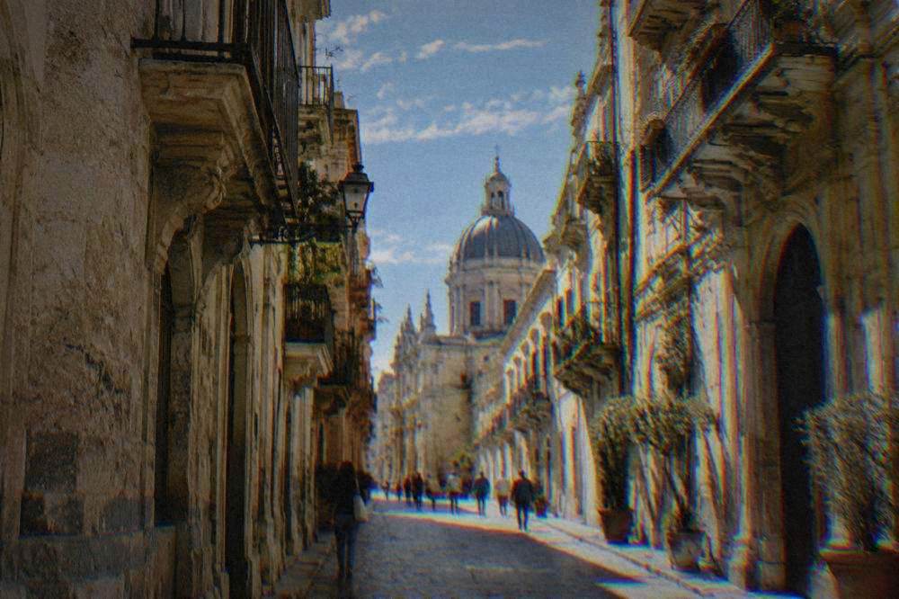 A busy street between golden limestone palazzi with balconies and a church dome rising beyond