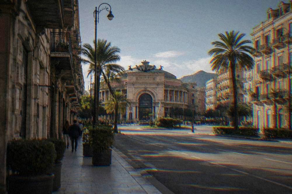 A broad boulevard of balconied apartment blocks and palm trees leading to a round piazza with an arched theatre facade