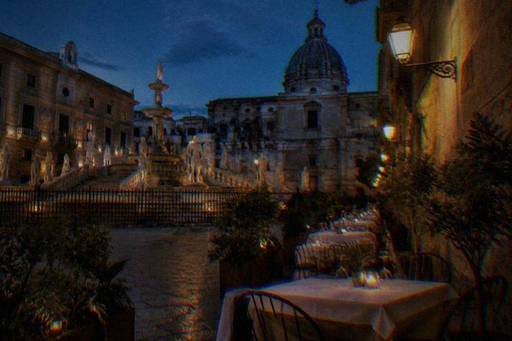 Outdoor restaurant tables with white cloths beside a stone wall in a lit lane at dusk