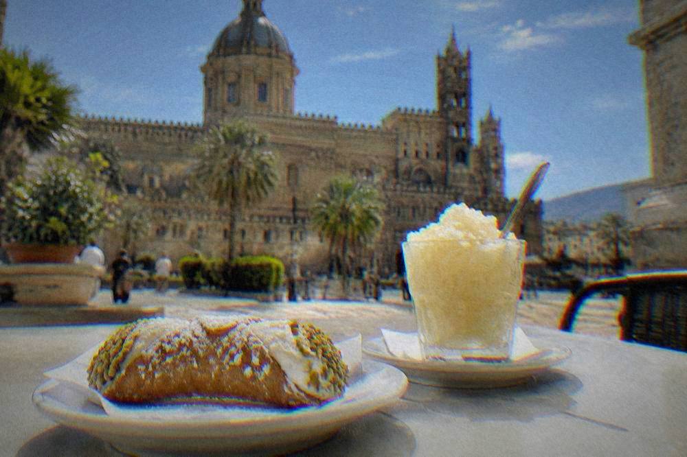 A café table with a ricotta pastry and a glass of granita in a sunny piazza