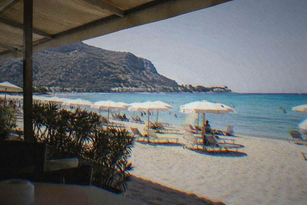 A wide crescent bay of pale sand and turquoise water with rows of plain parasols below a rocky headland