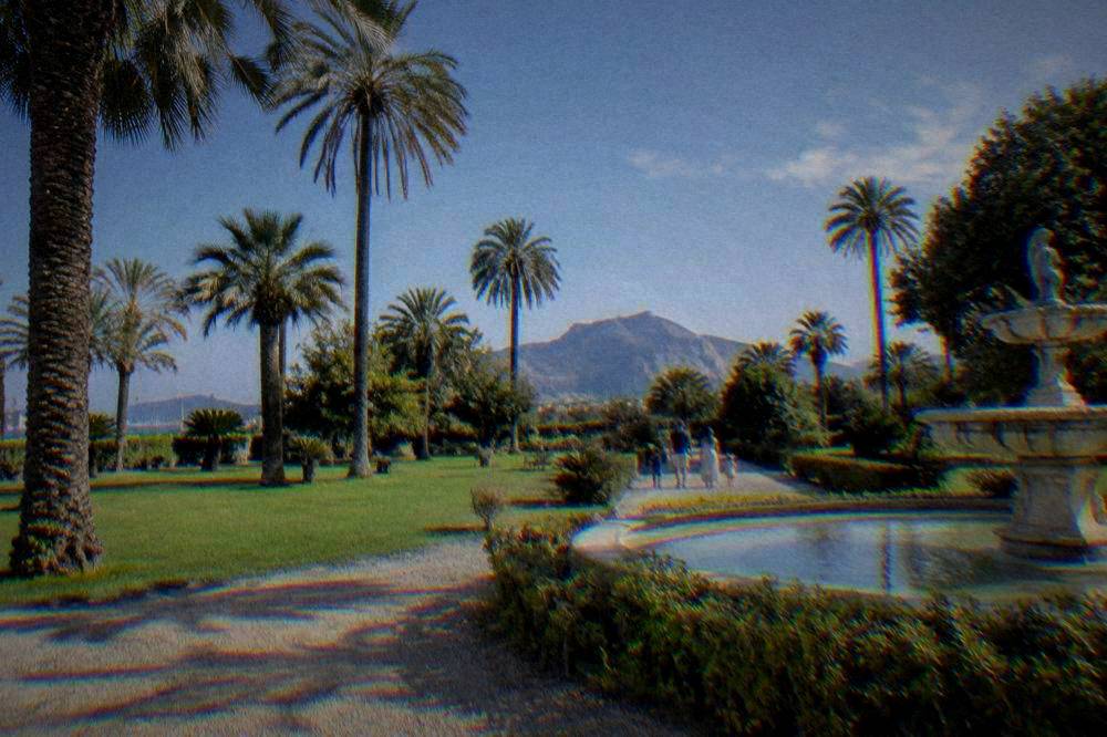 A sunny garden lawn with a gravel path, palms and a stone fountain with a family far off