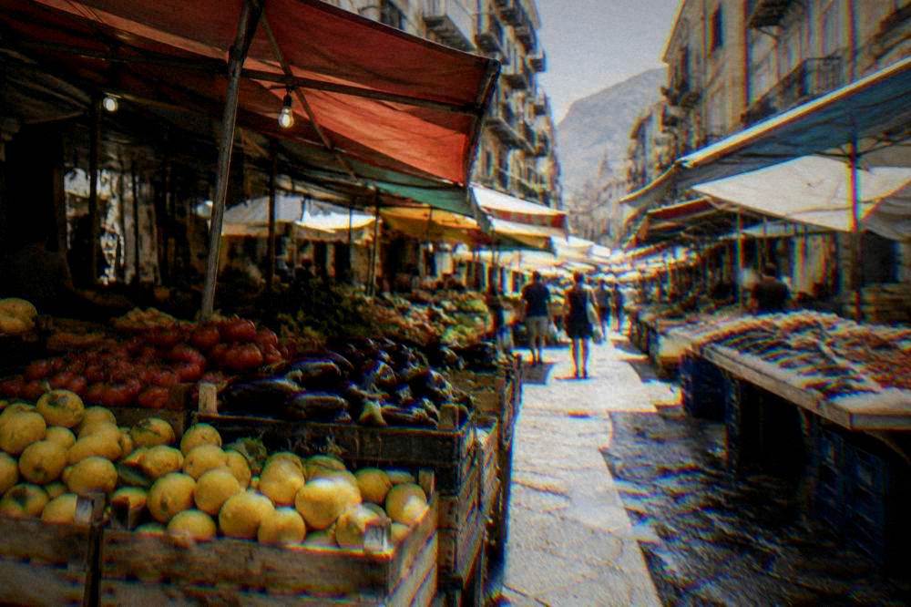 A market lane with stalls of lemons, tomatoes and aubergines and fish on ice below balconied facades