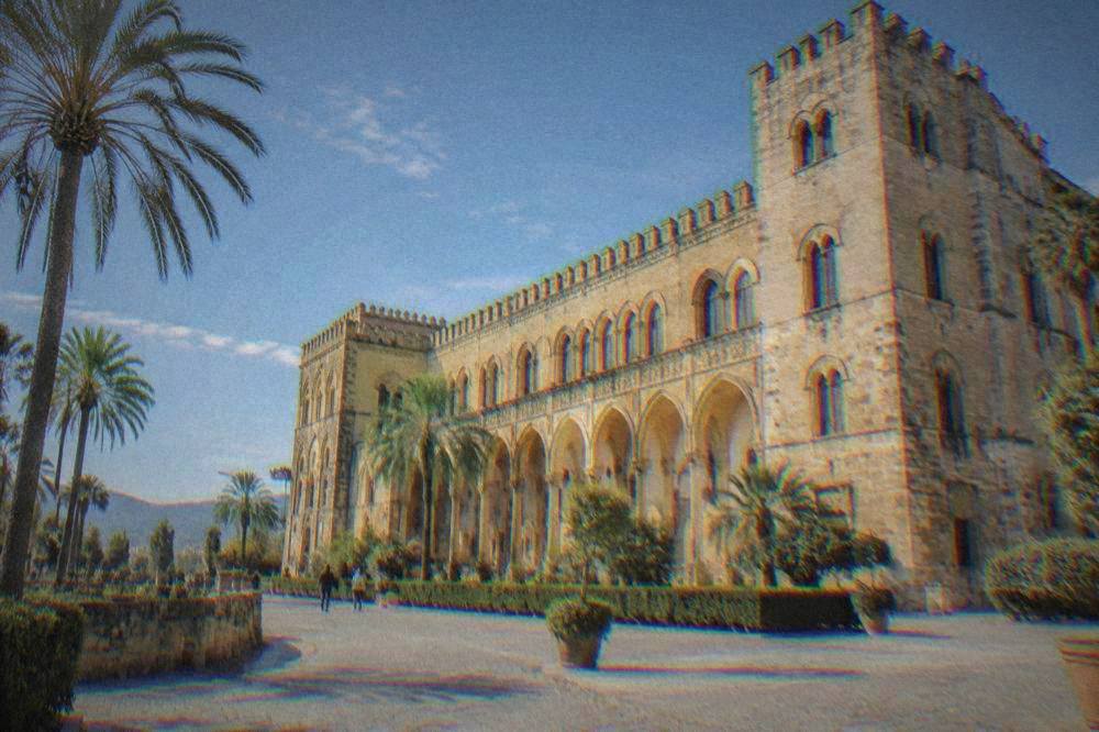 A long golden-stone palace facade with rows of arches and a crenellated tower behind palm trees