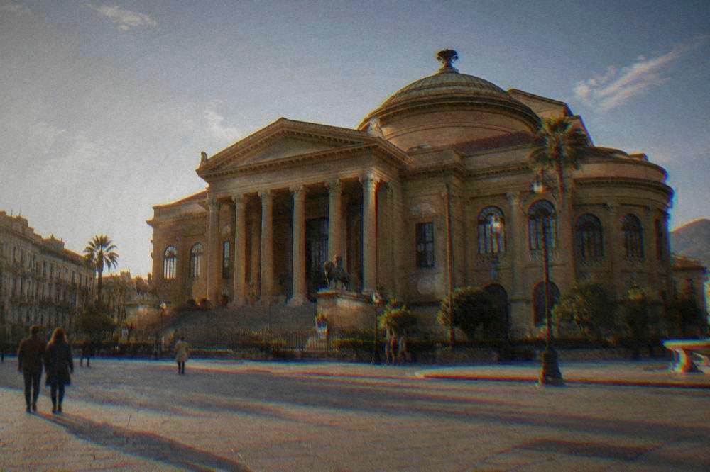A neoclassical opera house with a broad flight of steps, columned portico and green dome above a wide piazza