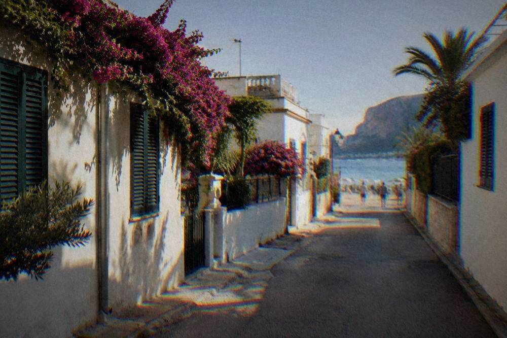 A quiet seaside street of low white villas with shutters and bougainvillea, the sea glimpsed at the end