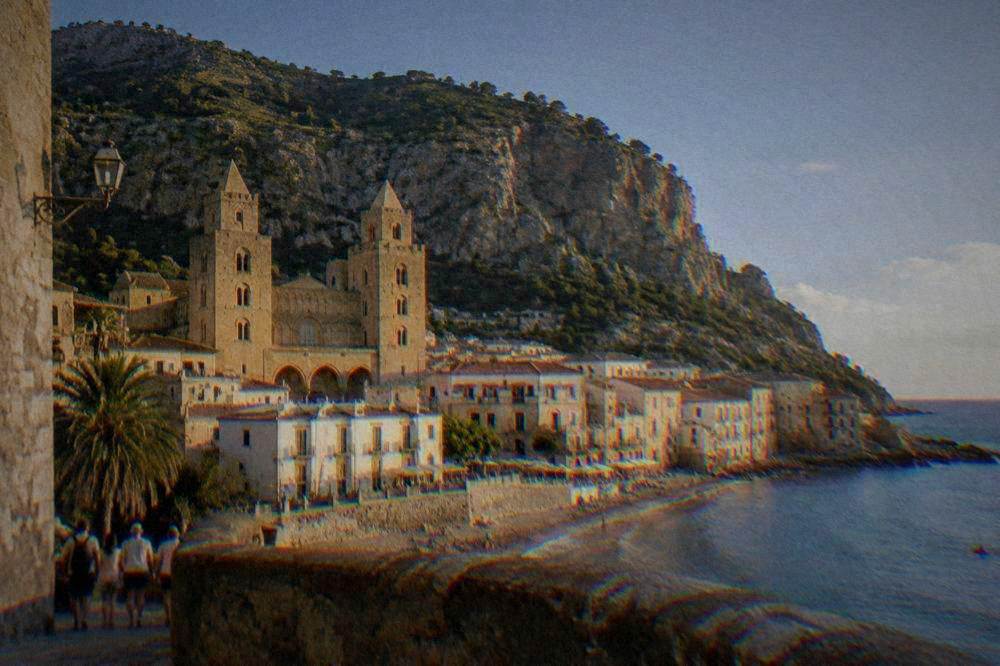 A seaside town with a twin-towered stone cathedral, terracotta roofs and a rocky headland behind a curve of beach