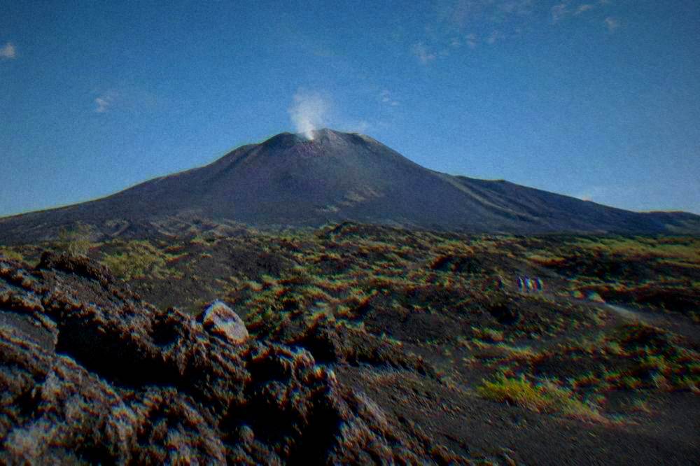 A broad black volcanic cone with a thin plume of steam above dark ash and lava rock
