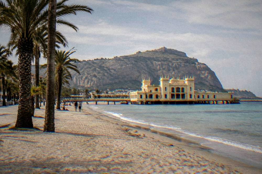 A wide sandy beach with palm trees and a period bathing pavilion at Mondello
