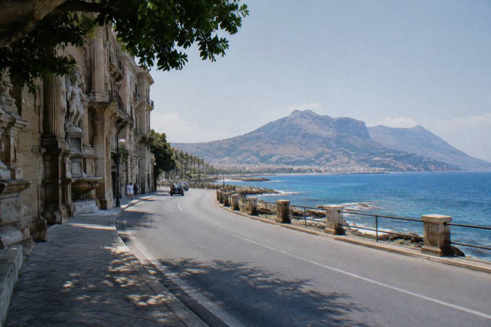 Coastal road curving along a blue sea toward distant Sicilian hills